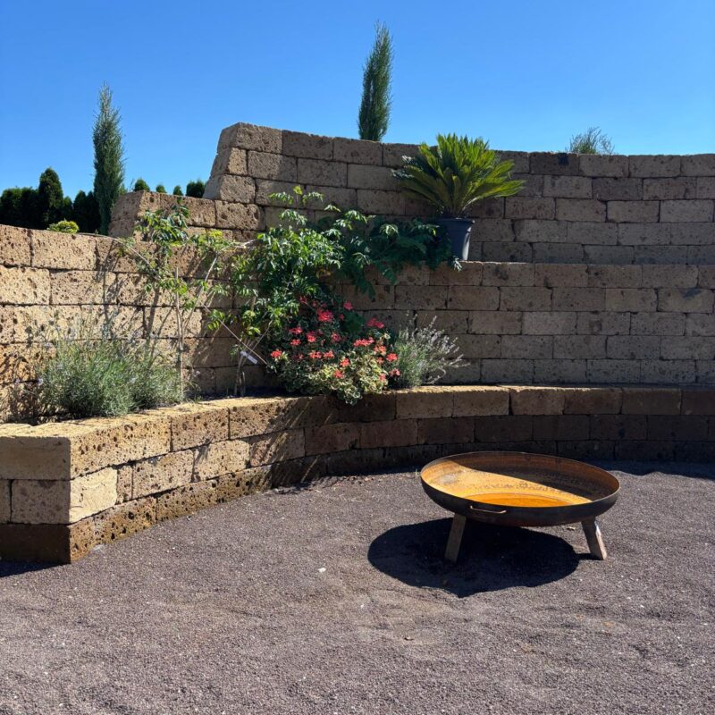 Feuerplatz mit runder Feuerschale auf Kiesfläche, umgeben von Natursteinmauern und mediterranen Pflanzen unter blauem Himmel.