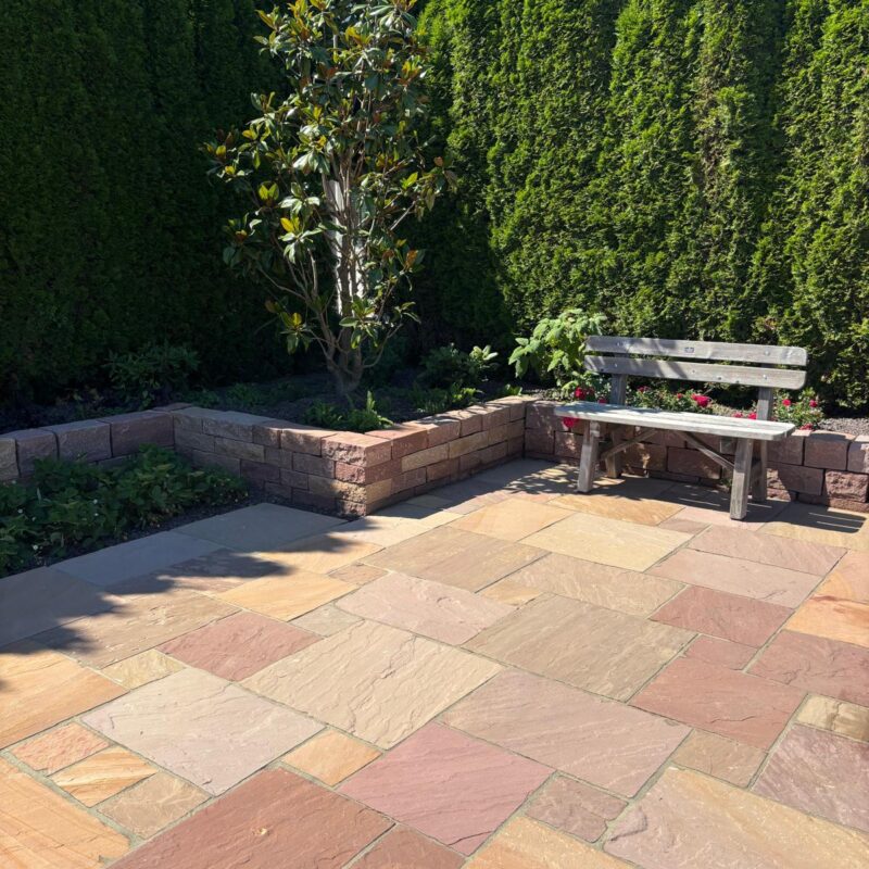 Zeigt rötlichen Natursteinbelag Red Sandstone als Terrasse mit Holzbank und Bepflanzung
